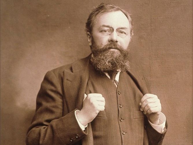 Photographie sépia, portrait en plan américain d'André Michelin vers 1890. Il adopte une posture fière, tenant de part et d'autre le veston qu'il porte sous son manteau.