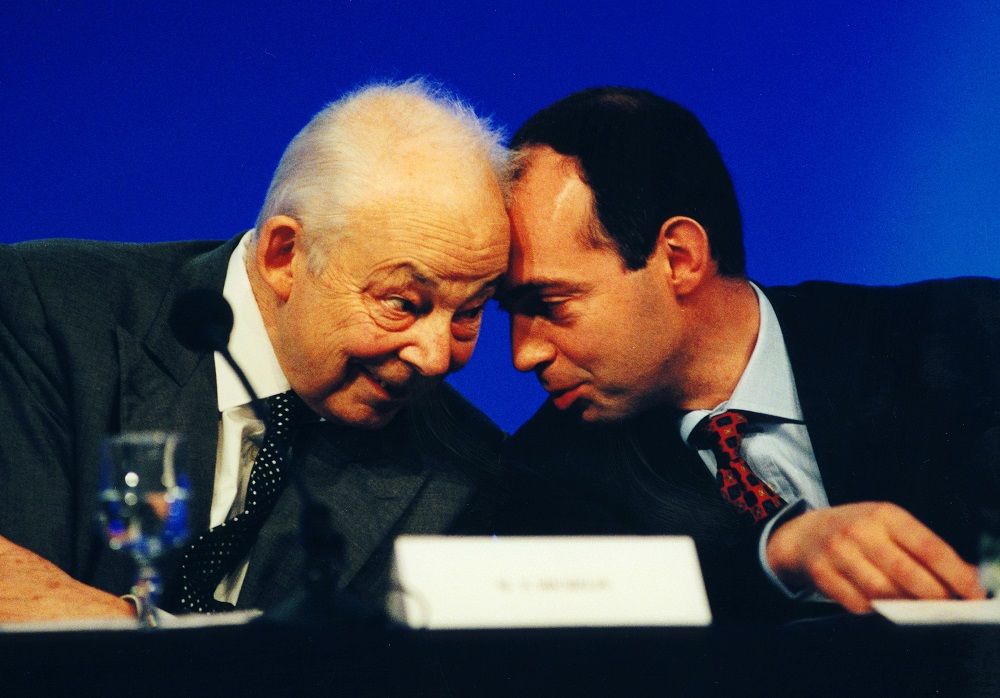 Photographie : portrait en plan rapproché de François Michelin (à gauche) et de son fils Édouard (à droite). Les deux hommes sont penchés l'un vers l'autre, leurs fronts se touchent tant ils sont proches. Ils échangent un regard complice.
